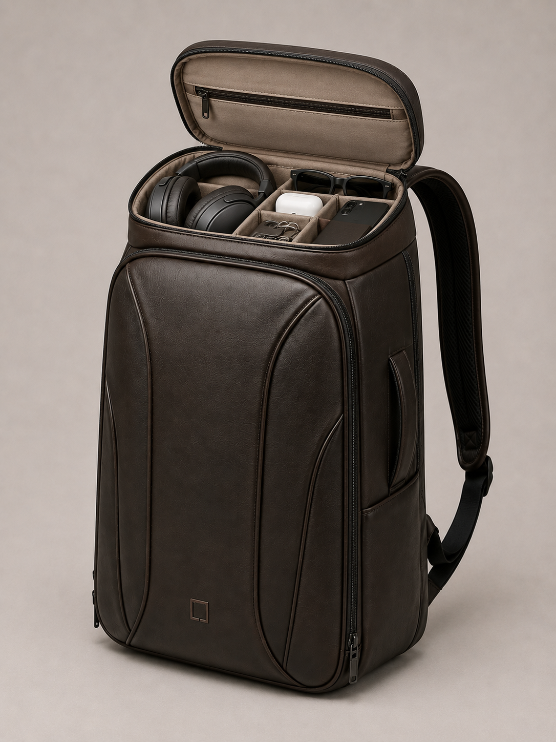A dark brown leather laptop backpack shown open, revealing a beige interior organizer with black headphones and other accessories inside. The backpack has a small square logo on the lower front and a black shoulder strap, photographed against a neutral beige background.
