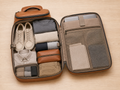 An open brown leather suitcase laid flat on a light wood surface, filled with neatly folded travel items including shoes, folded garments, a collared shirt, socks, and small zip pouches. The suitcase interior and compartments are visible, and no person or real environment beyond the tabletop setting is the focus.